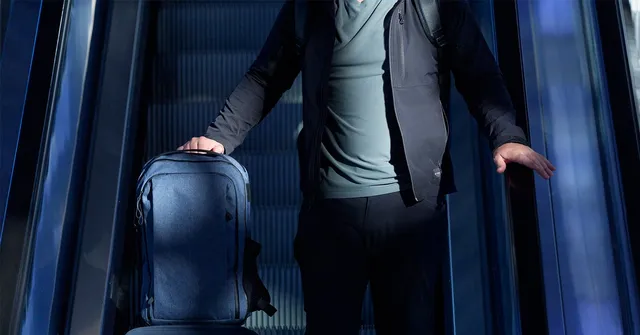 A person wearing dark clothing stands on an escalator, holding a blue suitcase with one hand and resting the other hand on the escalator railing. The…