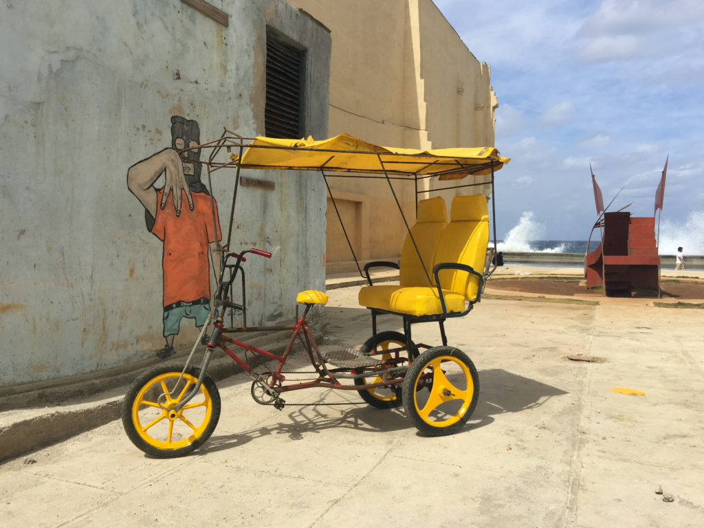 A freshly painted bike taxi sits in front of a graffiti mural as a wave crashes over the ocean wall on the Malecon. 